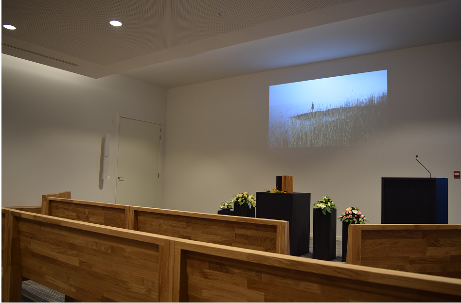 Aula met houten zitbanken, bloemstukken, lessenaar en projectie