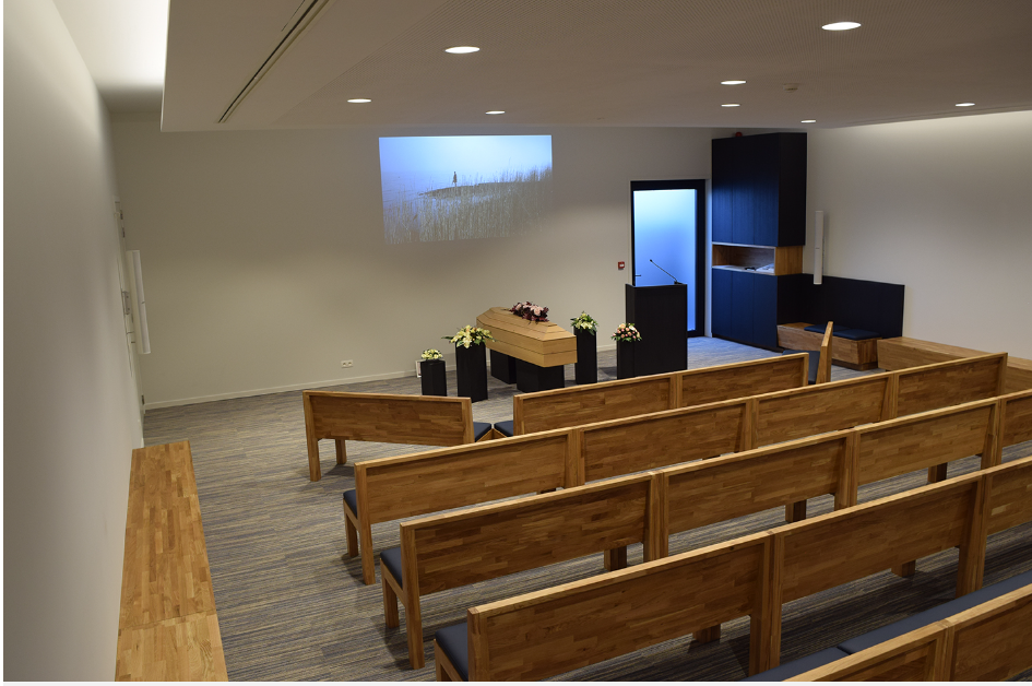 Aula van bovenaf met houten zitbanken, kist en bloemen
